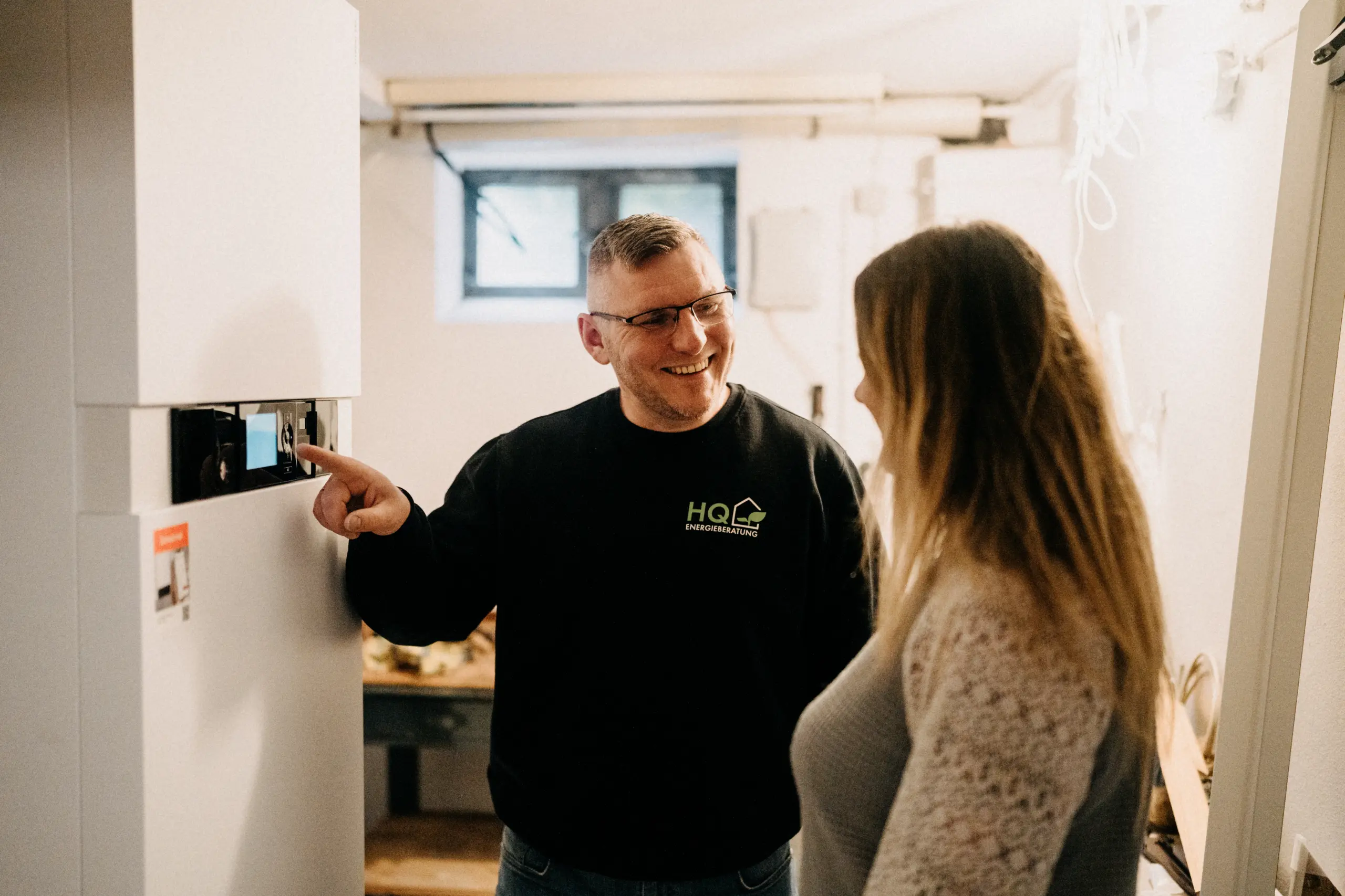 Florian Handschuh von HQ Energieberatung erklärt einer Kundin die Heizungsanlage im Heizungskeller bei einem Vor-Ort-Termin