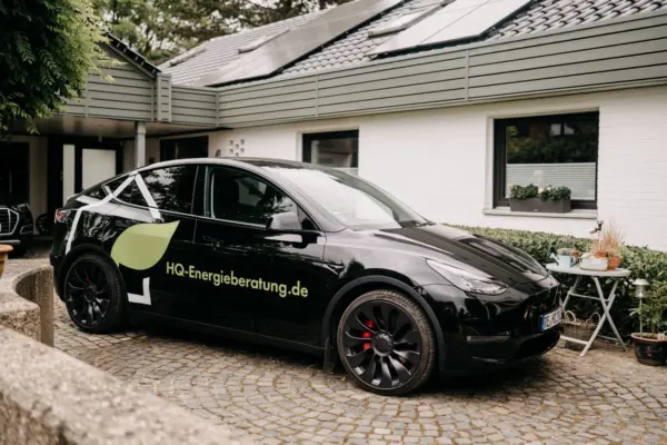 Schwarzer Tesla von HQ Energieberatung mit Logo und Webadresse vor einem Wohnhaus mit Solaranlage auf dem Dach